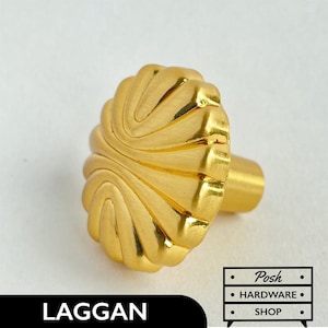 Pode incluir: Botão de armário dourado com um design em forma de concha com babados. O botão tem uma base redonda e um caule cilíndrico curto.