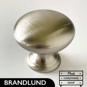 Może przedstawiać: Gałka meblowa z szczotkowanego niklu z okrągłym, lekko wypukłym wierzchołkiem i mniejszą, okrągłą podstawą. Gałka jest wykonana z metalu i ma gładkie, polerowane wykończenie.