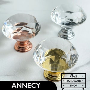 Könnte beinhalten: Drei Kristall-Schrankknöpfe mit verschiedenen farbigen Sockeln. Die Knöpfe sind diamantförmig und haben ein facettierendes Design. Die Sockel sind golden, silbern und kupferfarben.