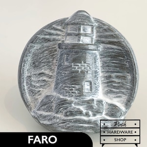May include: A white and grey ceramic lighthouse knob with a weathered finish. The knob is round and has a raised design of a lighthouse on a rocky shore. The text "FARO" is printed below the knob.