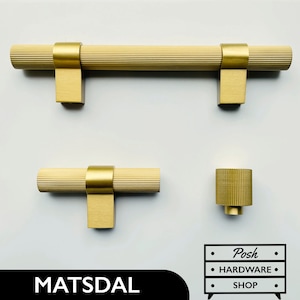 Pode incluir: Três botões de armário dourados com um design canelado. Os botões são de tamanhos diferentes. O botão maior é uma puxador. O botão menor é um botão redondo. O botão do meio é um botão em forma de T. O texto "MATSDAL" e "Posh Hardware Shop" é visível na imagem.