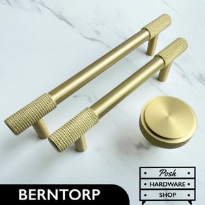 Peut inclure: Deux poignées de porte en or texturées avec un motif strié. Les poignées mesurent 25,4 cm de long et ont une forme en T. Un bouton assorti en or est également présenté. Le texte "BERNTORP" et "Posh Hardware Shop" est visible sur l'image.