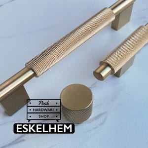 May include: Two gold-toned metal cabinet pulls and one matching round cabinet knob, all featuring a textured knurled surface. The hardware is displayed with a logo graphic that reads Posh HARDWARE SHOP and the text ESKELHEM in a black box.