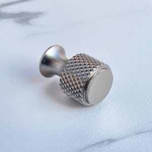 May include: A silver-toned cabinet knob with a textured, diamond-patterned grip and a smooth, circular end. The knob has a cylindrical shape with a flared base and is designed for easy handling. This hardware is suitable for drawers and cabinets.