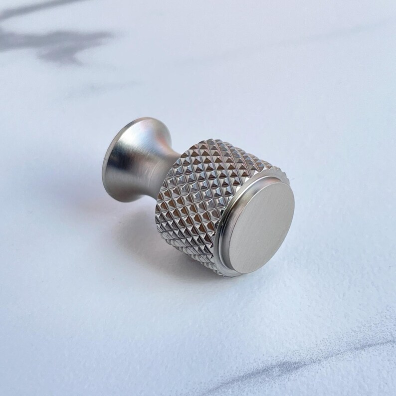 May include: A silver-toned cabinet knob with a textured, diamond-patterned grip and a smooth, circular end. The knob has a cylindrical shape with a flared base and is designed for easy handling. This hardware is suitable for drawers and cabinets.