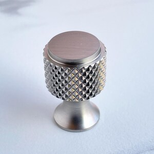 May include: A silver-toned cabinet knob with a diamond-patterned grip. The knob has a cylindrical body with a flat top and a flared base. The metal hardware is designed for furniture and home decor.