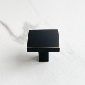 Op de afbeelding: Zwarte vierkante kastknop met een gouden rand en een zwarte pen.