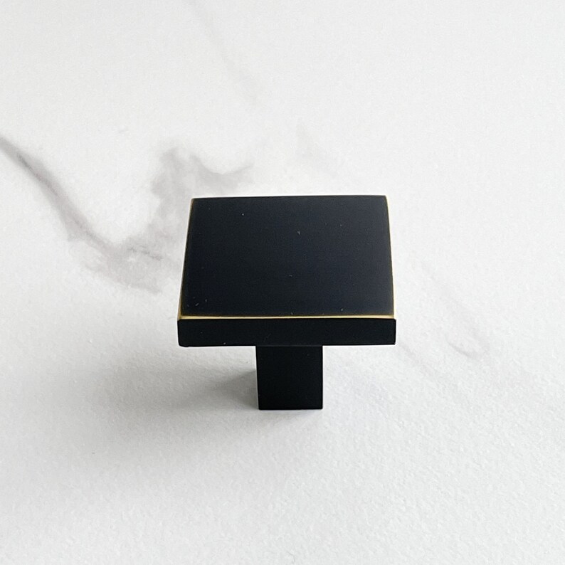 Op de afbeelding: Zwarte vierkante kastknop met een gouden rand en een zwarte pen.