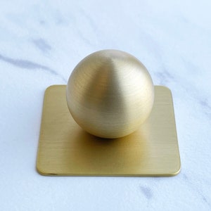 May include: A gold-colored metal sphere sits on a square, gold-colored metal base. Both the sphere and the base have a brushed finish.