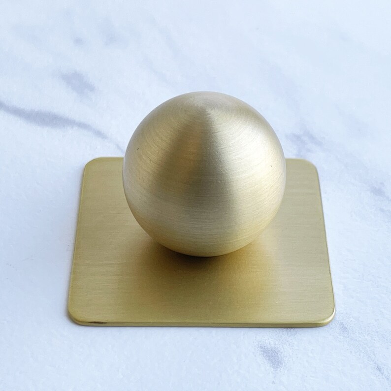 May include: A gold-colored metal sphere sits on a square, gold-colored metal base. Both the sphere and the base have a brushed finish.