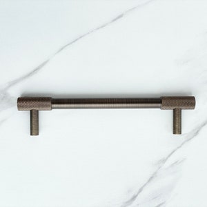 May include: A dark bronze cabinet pull with a cylindrical bar and two vertical mounting posts. The ends of the bar feature a textured, cross-hatched knurled pattern, while the center of the bar and the mounting posts have a smooth, brushed finish.