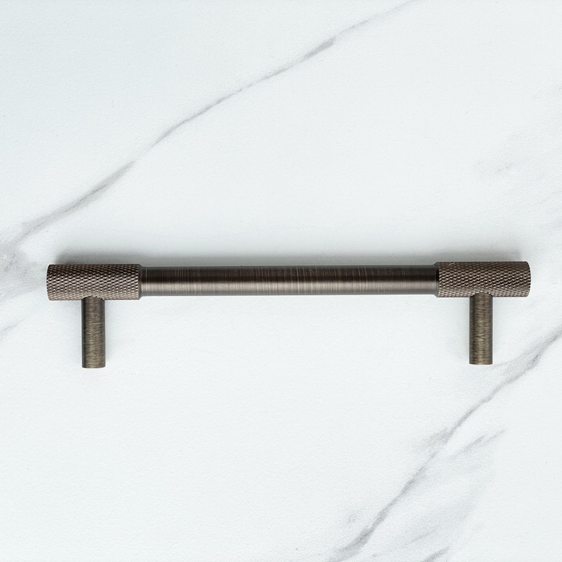 May include: A dark bronze cabinet pull with a cylindrical bar and two vertical mounting posts. The ends of the bar feature a textured, cross-hatched knurled pattern, while the center of the bar and the mounting posts have a smooth, brushed finish.