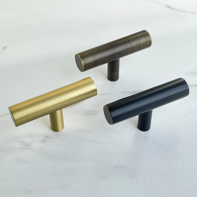 K&ouml;nnte beinhalten: Drei Schrankkn&ouml;pfe in verschiedenen Ausf&uuml;hrungen: Gold, Messing und Schwarz. Jeder Knopf hat eine zylindrische Form mit einem T-f&ouml;rmigen Griff.