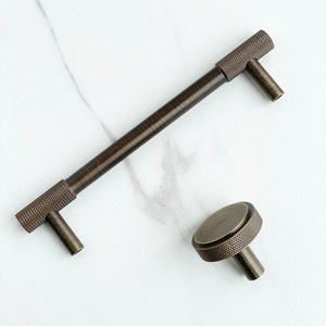 May include: A dark bronze cabinet handle and a matching round cabinet knob. Both pieces feature a textured, cross-hatched knurled pattern on the cylindrical sections and a smooth, brushed metallic finish on the flat surfaces.