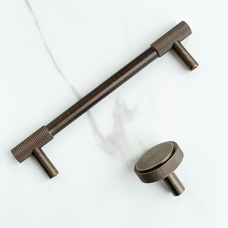 May include: A dark bronze cabinet handle and a matching round cabinet knob. Both pieces feature a textured, cross-hatched knurled pattern on the cylindrical sections and a smooth, brushed metallic finish on the flat surfaces.