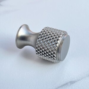 May include: A silver-toned cabinet knob with a diamond-patterned grip. The knob has a cylindrical body and a flared base, suitable for furniture or decorative applications. The overall design is modern and minimalist.