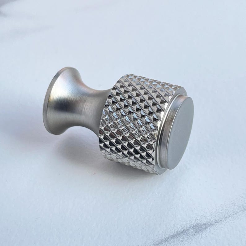 May include: A silver-toned cabinet knob with a diamond-patterned grip. The knob has a cylindrical body and a flared base, suitable for furniture or decorative applications. The overall design is modern and minimalist.