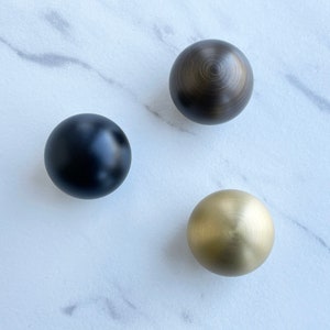 May include: Three round cabinet knobs in different finishes: black, gold, and bronze. The knobs are on a white marble surface.