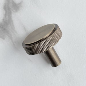 May include: A round cabinet knob with an antique brass finish. The knob features a flat circular top surface and a textured, diamond-patterned knurled edge around the circumference. A cylindrical stem extends from the center of the base.