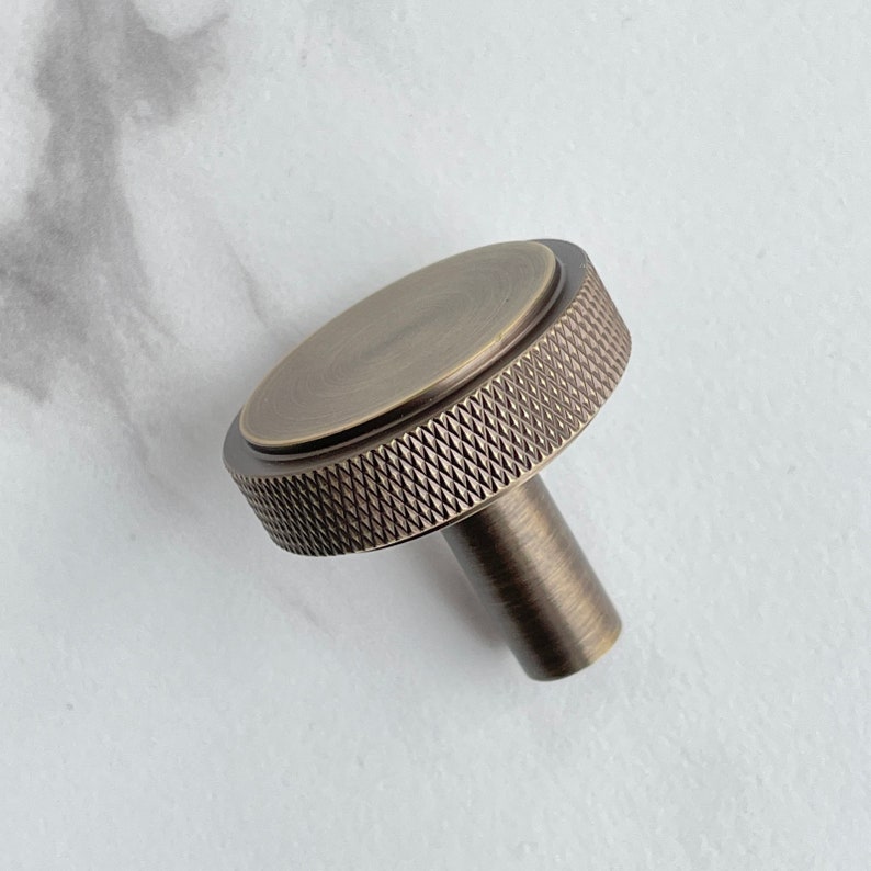 May include: A round cabinet knob with an antique brass finish. The knob features a flat circular top surface and a textured, diamond-patterned knurled edge around the circumference. A cylindrical stem extends from the center of the base.