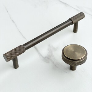 May include: A dark bronze cabinet handle and a matching round cabinet knob. The handle features a long, smooth central bar with knurled textured ends. The round knob has a flat, circular top surface with a knurled edge and a central post.