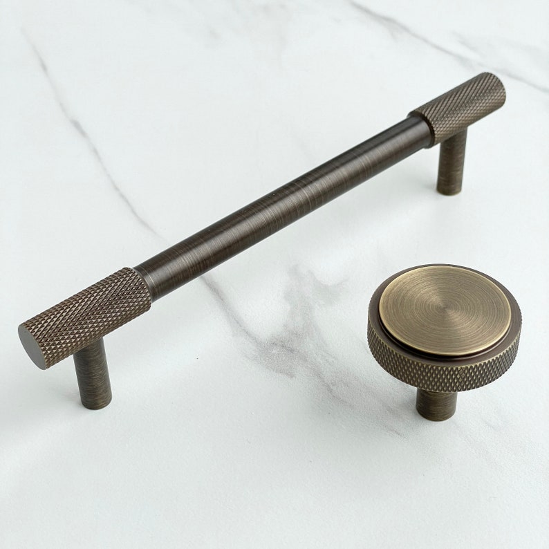 May include: A dark bronze cabinet handle and a matching round cabinet knob. The handle features a long, smooth central bar with knurled textured ends. The round knob has a flat, circular top surface with a knurled edge and a central post.
