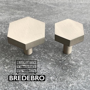 May include: Two hexagonal cabinet knobs with a brushed metallic finish. The knobs feature a flat, six-sided top surface and a cylindrical base. A logo graphic is visible in the lower left corner with the text Posh Hardware Shop Bredebro.