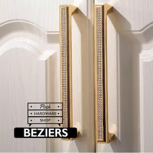 May include: Two vertical gold cabinet handles featuring a central strip of embedded clear crystals. The handles are mounted on white cabinet doors. A logo with the text Posh HARDWARE SHOP and the word BEZIERS is visible in the lower left corner.