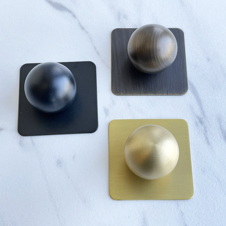 May include: Three round knobs in different finishes: black, brass, and gold. Each knob is mounted on a square base.