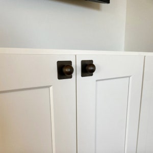 May include: Two white cabinet doors with square bronze knobs. The doors are closed and have a raised panel design.