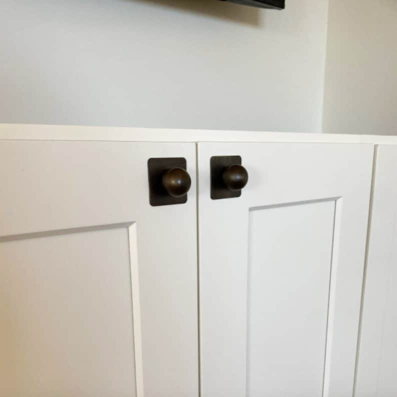 May include: Two white cabinet doors with square bronze knobs. The doors are closed and have a raised panel design.