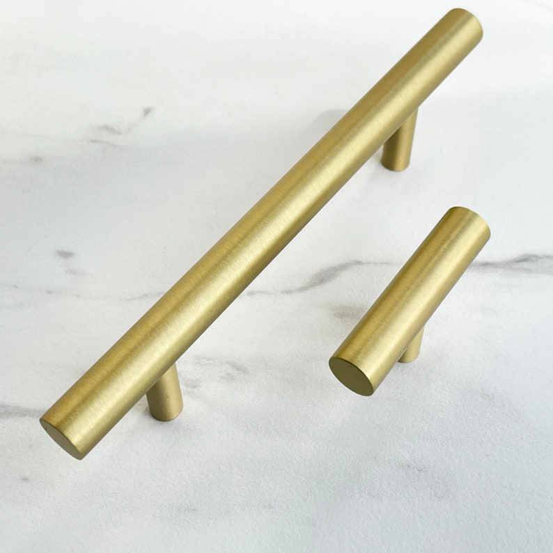 K&ouml;nnte beinhalten: Zwei geb&uuml;rstete goldene Schrankgriffe, einer lang und einer kurz, auf einer wei&szlig;en Marmorfl&auml;che.