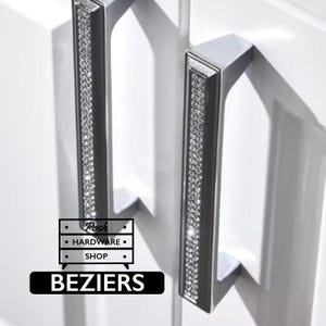 May include: Two rectangular silver cabinet pulls with a vertical inlay of clear crystals. The handles are mounted on white cabinet doors. A logo graphic is visible in the lower left corner with the text Posh HARDWARE SHOP and BEZIERS.