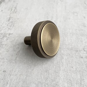 May include: A round cabinet knob with a brushed brass finish on the front face and a dark bronze-toned knurled edge. The knob features a central circular plate with a radial brushed texture, surrounded by a textured, cross-hatched grip pattern.
