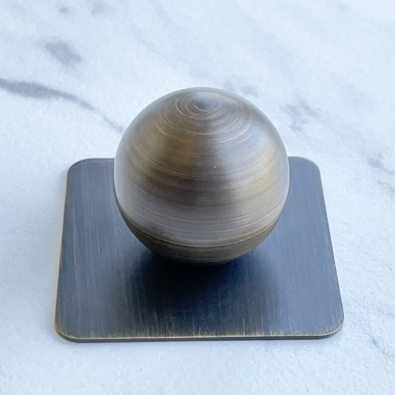 May include: A round, brushed brass knob with a square, brushed brass base.