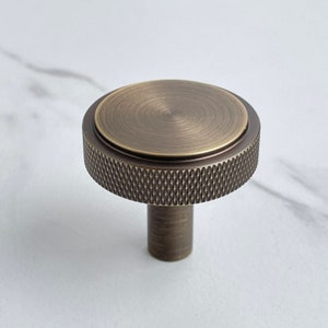 May include: A round cabinet knob with a brushed antique brass finish. The top surface is smooth and circular, while the outer edge features a textured, diamond-patterned knurled grip. A cylindrical base extends downward from the center.