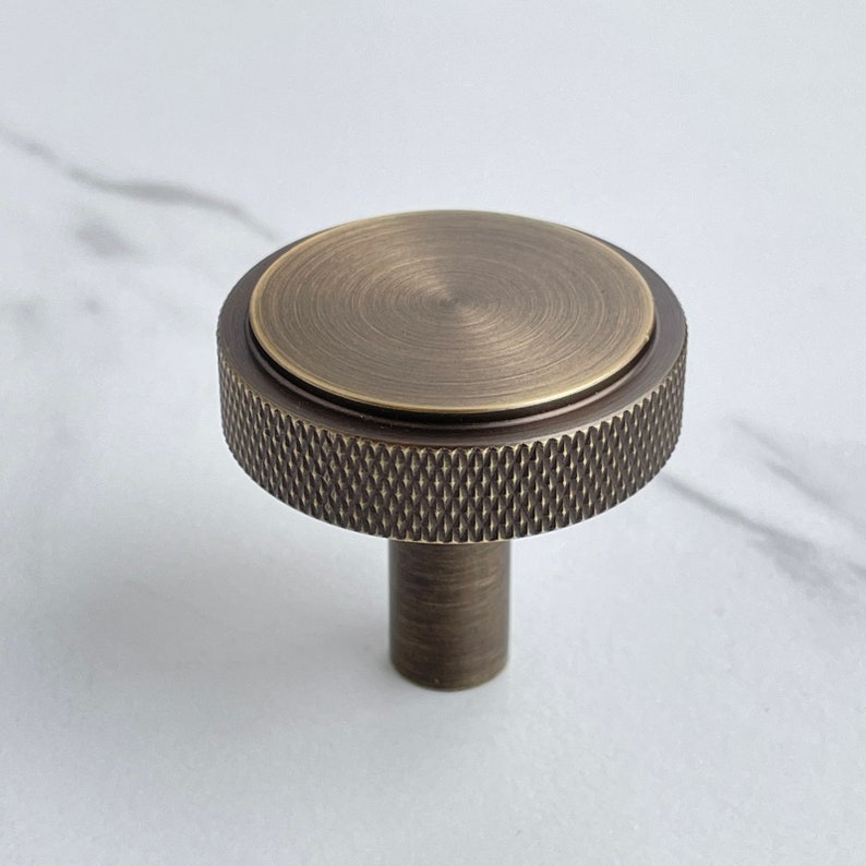 May include: A round cabinet knob with a brushed antique brass finish. The top surface is smooth and circular, while the outer edge features a textured, diamond-patterned knurled grip. A cylindrical base extends downward from the center.