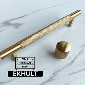 Può includere: Una maniglia per mobili in metallo dorato con una sezione centrale testurizzata e zigrinata e due estremità cilindriche lisce, accompagnata da un pomello circolare abbinato. L'immagine include un logo con il testo Posh Hardware Shop e EKHULT.