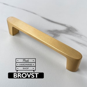 May include: A brushed gold cabinet handle with a smooth, elongated rectangular bar and rounded ends. The hardware features a matte metallic finish. A logo graphic is visible in the corner with the text Posh HARDWARE SHOP and BROVST.