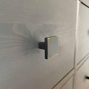 Op de afbeelding: Een zwarte vierkante kastknop met een gouden rand. De knop is gemonteerd op een grijze houten lade.