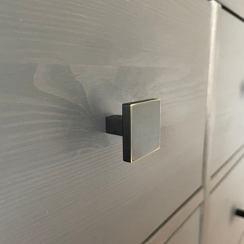 Op de afbeelding: Een zwarte vierkante kastknop met een gouden rand. De knop is gemonteerd op een grijze houten lade.