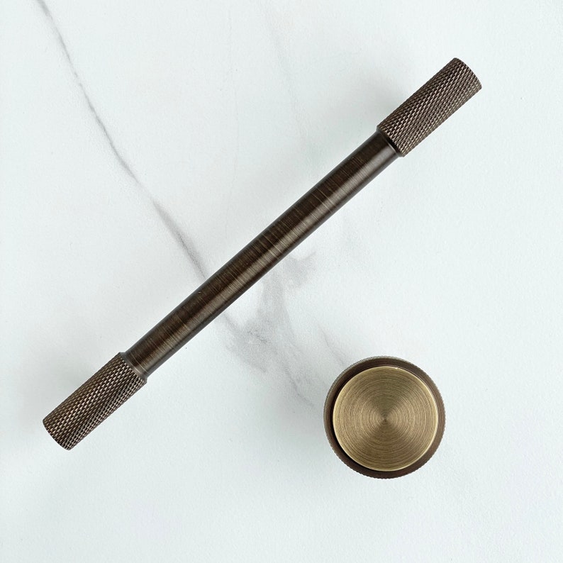 May include: A dark bronze-toned metal cabinet handle with a long, smooth cylindrical center and textured, diamond-patterned knurled ends, accompanied by a matching circular cabinet knob featuring a brushed brass-toned flat top surface.