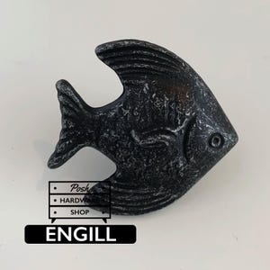 Può includere: Un pomello per mobili a forma di pesce con una finitura grigio antracite scuro e una texture usurata e invecchiata. La superficie presenta dettagli in rilievo di pinne e squame. Sotto il pesce, il logo Posh HARDWARE SHOP e il testo ENGILL.