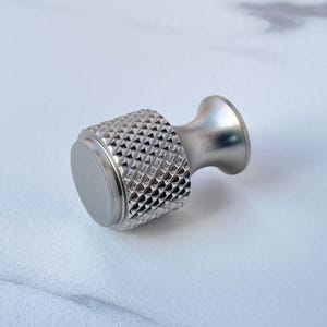 May include: A silver-toned cabinet knob with a diamond-patterned grip and a smooth, circular base. The knob has a flared, rounded handle. The hardware is made of metal and is designed for use on drawers or cabinets.