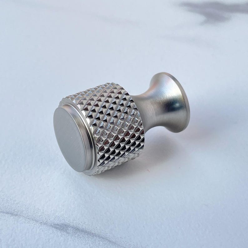 May include: A silver-toned cabinet knob with a diamond-patterned grip and a smooth, circular base. The knob has a flared, rounded handle. The hardware is made of metal and is designed for use on drawers or cabinets.