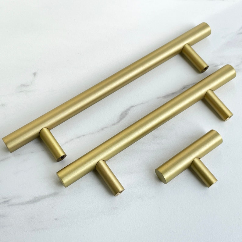 K&ouml;nnte beinhalten: Drei goldfarbene Schrankgriffe in verschiedenen Gr&ouml;&szlig;en. Die Griffe haben eine T-Form mit einem geraden Balken und einem senkrechten Griff.