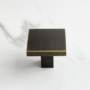 Op de afbeelding: Een vierkante kastknop met een geborsteld messing afwerking en een zwarte basis.