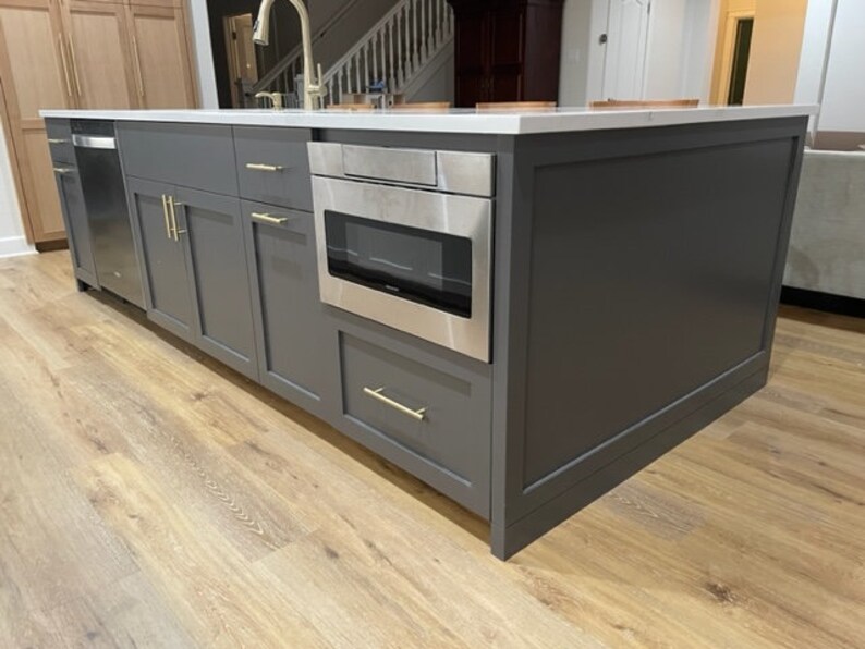 K&ouml;nnte beinhalten: Eine dunkelgraue K&uuml;cheninsel mit einer wei&szlig;en Arbeitsplatte und einem eingebauten Edelstahlbackofen. Die Insel hat goldene Beschl&auml;ge und steht auf einem hellen Holzboden.