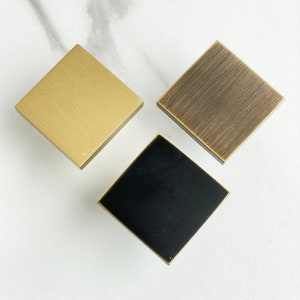Op de afbeelding: Drie vierkante kastknoppen in verschillende afwerkingen: goud, geborsteld messing en zwart.