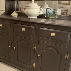 May include: A dark brown wooden hutch with three drawers and two doors. The hutch has brass hardware and a mirrored back.